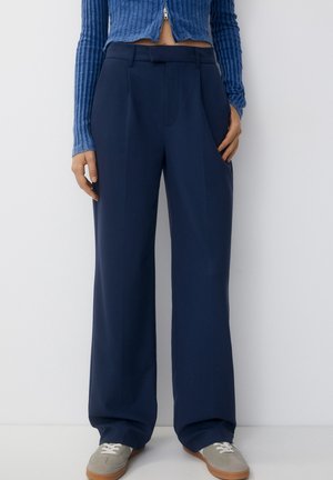 Person trägt marineblaue Hosen mit weitem Bein, graue Sneakers und einen gerippten blauen Cropped-Pullover mit Frontreißverschluss, steht vor weißem Hintergrund.