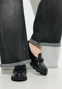 Zwarte leren loafers met een kettingaccent, met een chunky zool. Draagt men met donkere spijkerbroeken, opgerold aan de mouwen, die subtiele details tonen.
