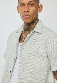 Chemise à manches courtes avec boutons en tissu texturé gris et blanc, dotée d'un col évasé et de boutons noirs. Portée par-dessus un débardeur blanc.