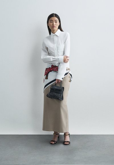 Mulher com uma camisa branca de botões, saia longa bege com botões laterais, sandálias pretas, segurando uma pequena bolsa preta com um lenço estampado atado à cintura.