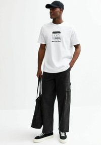 Camiseta de algodón blanca con un estampado gráfico y letras negras, combinada con pantalones cargo de mezclilla negra holgados y zapatillas negras. Llevando una bolsa negra.