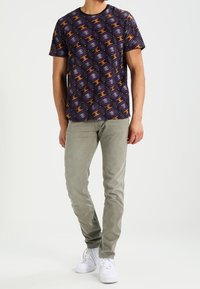 Fioletowa koszula z krótkim rękawem z abstrakcyjnym pomarańczowo-żółtym wzorem, zestawiona z jasnoszarymi dżinsami i białymi trampkami.