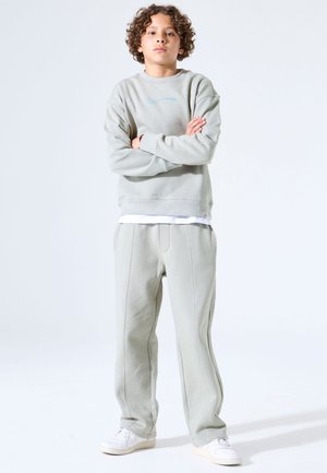 Junge Person steht mit verschränkten Armen, trägt hellgrauen Pullover, passende Hose und weiße Sneakers, vor schlichtem weißem Hintergrund.