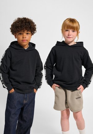 Zwarte hoodies met witte logo-accenten op de mouwen, gedragen door twee kinderen. De een combineert met denim, de ander met beige shorts.