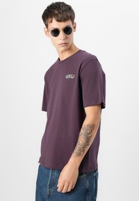 Joven con gafas de sol redondas negras, camiseta morada "Spell Bound", jeans azules y un tatuaje de un reloj y rosas en su antebrazo.