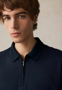 Marinblå zip-up polo med en texturerad krage och en figurnära design. Slät tyg och en diskret dragkedja i fram.