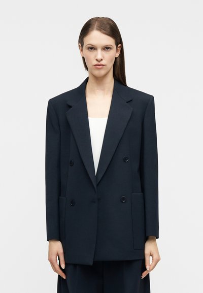 Femme aux cheveux bruns lisses portant un blazer noir surdimensionné à double boutonnage par-dessus un haut blanc, debout devant un fond clair uni.