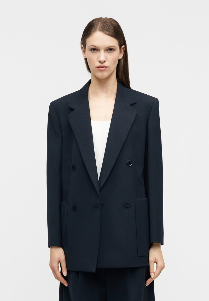 Γυναίκα με ίσια καστανά μαλλιά φορ άει υπερμεγέθη μαύρο διπλόμπουκλωτο σακάκι πάνω από λευκό τοπ, στέκεται μπροστά σε απλό φωτεινό φόντο.