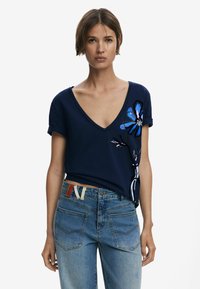 Marinblå V-ringad t-shirt med rullade ärmar, med en blå blommönstring på vänster sida. Bärs med ljusblå jeans och ett flerfärgat bälte.