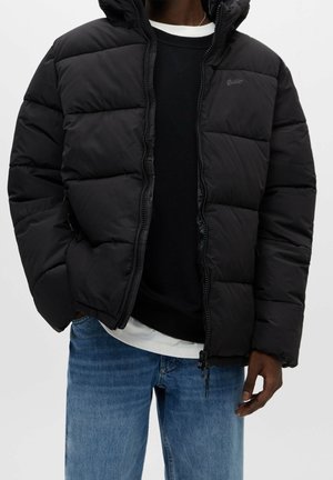 Man i svart pufferjacka över svarta och vita lagerade toppar med blå jeans, händerna avslappnat vid sidorna, mot en enfärgad bakgrund.
