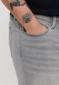 Sive traperice slim fit kroja, s prednjim džepom i metalnim detaljima. Pozadina je jednostavna, ističući teksturu materijala.
