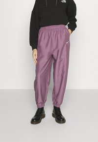 Person som bär lila Nike-joggers, svart North Face-tröja och svarta snörkängor, stående med en hand i fickan.