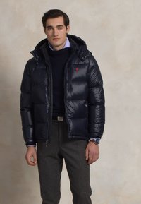 Diepblauwe pufferjack met capuchon, met een rits aan de voorkant en een gewatteerde structuur. Gedragen over een donkere trui en grijze broek.