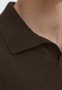 Gros plan sur le col d'un polo en tricot marron porté par une personne, montrant l'encolure et la partie supérieure de la poitrine.