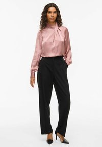 Blusa de seda rosa con un cuello alto y detalle drapeado, combinada con pantalones negros ajustados y tacones de punta negros.