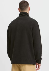 Black fleece pullover with a high collar, relaxed fit, and textured surface. Cuffed sleeves and a straight hem enhance functionality.