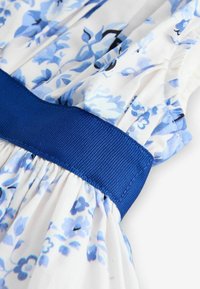 Balta medžiaga su mėlynu gėlių raštu ir klostuotomis detalėmis, su plačiu tekstūruotu tamsiai mėlynu juosmeniu.