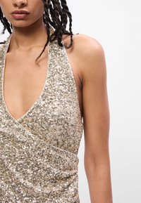 Χρυσό παγιέτα φόρεμα με λαιμόκοψη halter και βαθύ V, με σχέδιο περιτυλίγματος και λεία υφή, αναδεικνύοντας λαμπερές λεπτομέρειες.