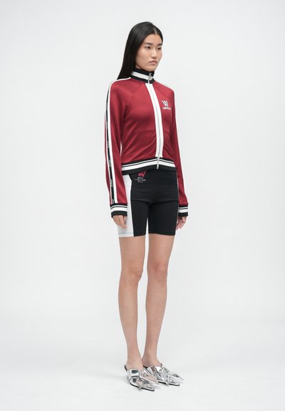 Giacca sportiva rossa con bordi bianchi, chiusura con zip e pantaloncini neri con dettagli grigi. Modello con scarpe slip-on argento metallizzato.