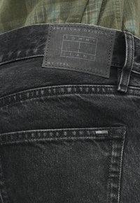 Buzunarul din spate și eticheta din piele a blugilor negri Tommy Jeans, cu cusături vizibile, și o parte din cămașa de catifea verde deasupra.