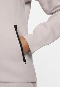 Licht mauve sweatshirt met een ritszak, geribde manchetten en een zachte textuur. Heeft een zwart ritsdetail voor contrast.