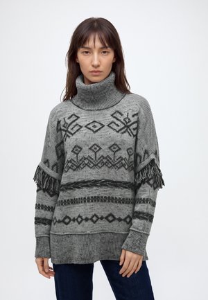 Femme portant un pull à col roulé en maille grise avec des motifs géométriques noirs et des franges aux manches, sur un fond uni.