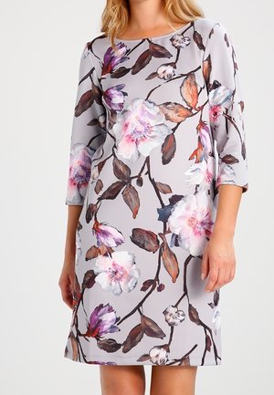 Femme portant une robe grise claire arrivant aux genoux, ornée de grandes imprimés floraux roses, blancs et violets, avec des manches trois-quarts.