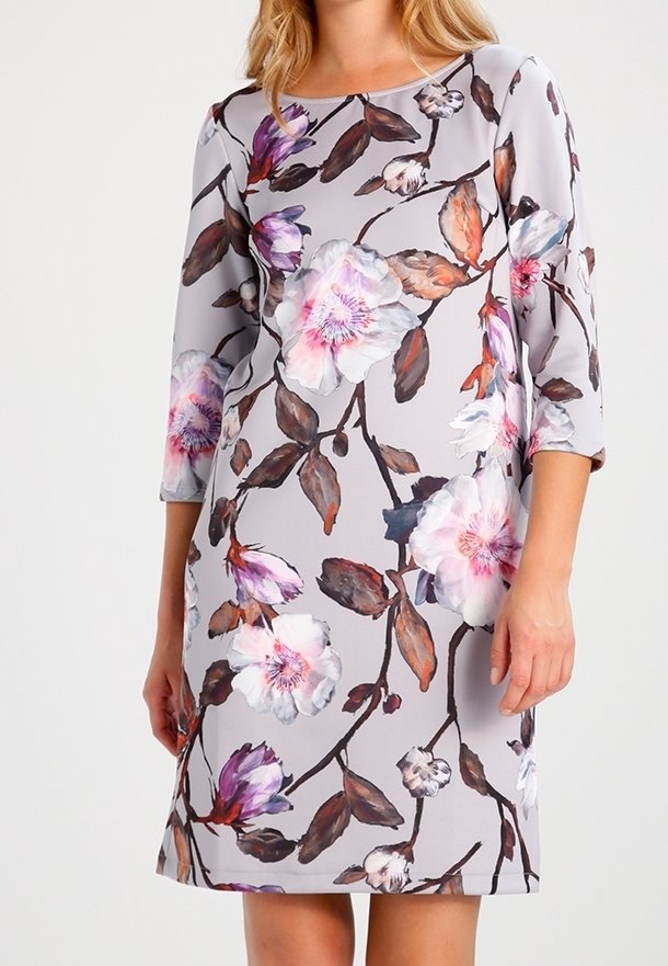 Kobieta ubrana w jasnoszarą sukienkę do kolan z dużymi różowymi, białymi i fioletowymi kwiatowymi wzorami oraz rękawami o długości do łokcia.
