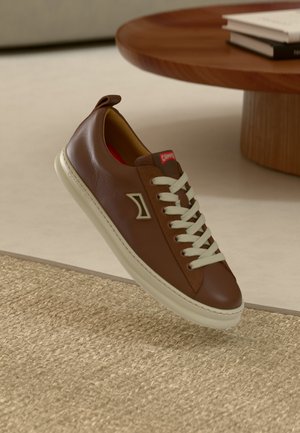 Brown leather sneaker with white laces, round toe, textured surface, and a red tag on the tongue. Cream-colored sole with a contrasting design.
