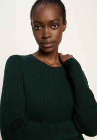 Junge Frau mit geflochtenem Haar, die einen dunkelgrünen gestreiften Pullover trägt und direkt in die Kamera mit einem neutralen Ausdruck schaut.