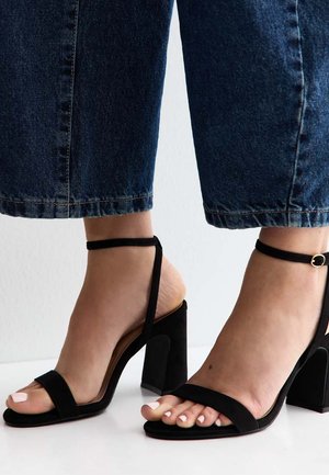Voeten met zwarte sandalen met blokhakken en enkelbandjes, gecombineerd met donkerblauwe cropped jeans tegen een effen witte achtergrond.