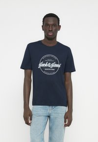 Tamnoplava pamučna majica s okruglim izrezom, s bijelim kružnim logotipom koji uključuje tekst. Nošena s svijetloplavim trapericama.