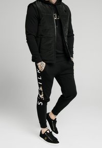 Uomo che indossa un gilet trapuntato nero, felpa con cappuccio nera, pantaloni jogger neri con testo "SikSilk" bianco e oro, e sneakers nere con suole bianche e dettagli dorati.