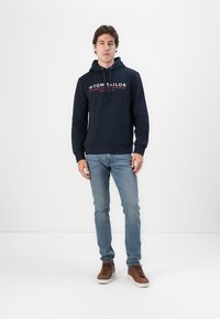 Granatowy hoodie z trokami i logo z przodu, zestawiony z jasnoniebieskimi dżinsami i brązowymi sneakersami. Gładki materiał i regularny krój.