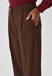 Pantaloni tailored marroni con una texture liscia, dotati di pieghe, tasche laterali e una cintura con un discreto particolare di chiusura.