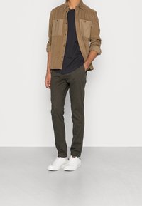 Bruine corduroy jas met twee borstzakken, zwarte t-shirt eronder, donkere broek en witte sneakers. Casual outfit gepresenteerd op een model.
