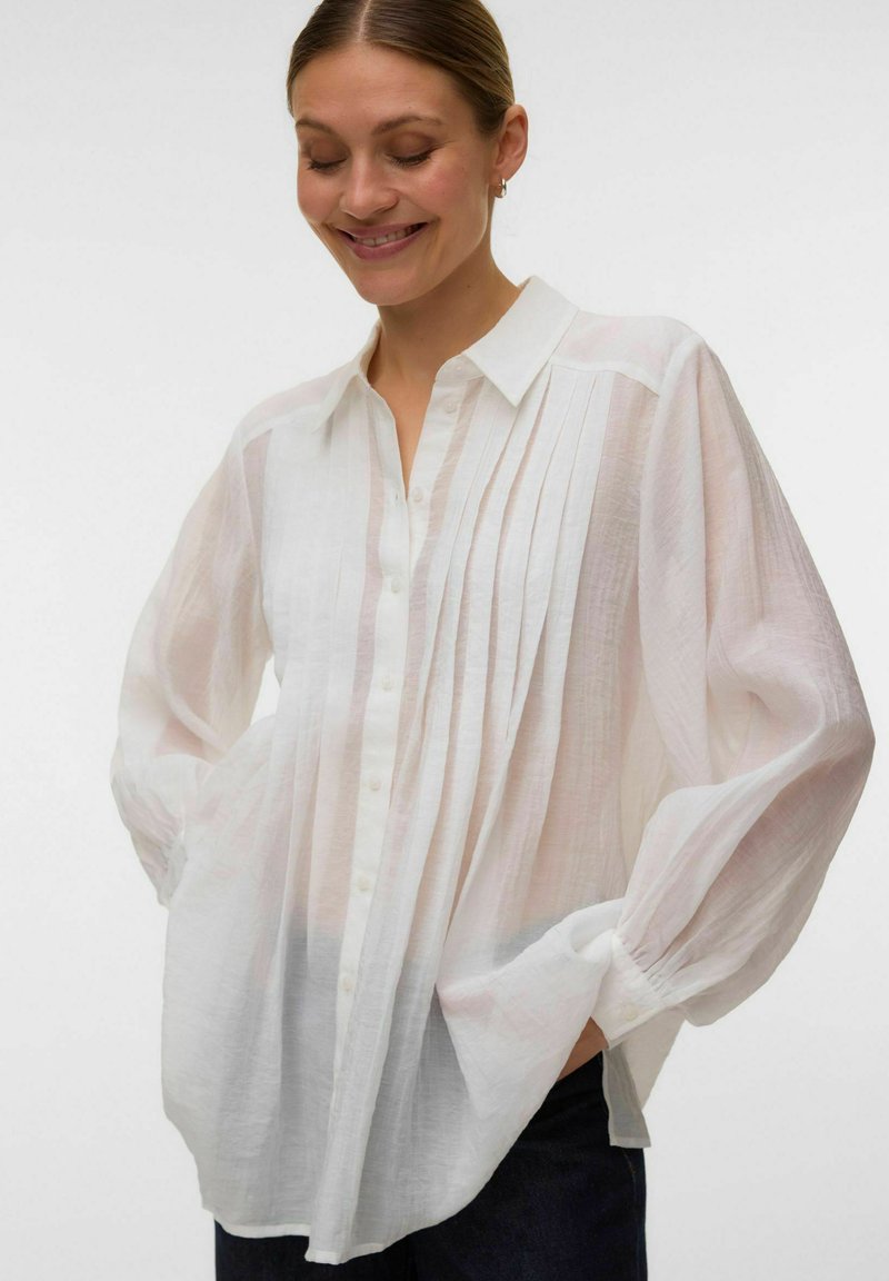 Femme souriante portant un chemisier blanc boutonné avec un devant plissé et des manches transparentes et amples, les mains dans les poches, sur un fond uni.