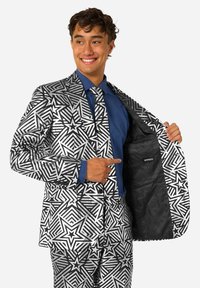 Chaqueta de traje con patrón de estrellas en blanco y negro con acabado brillante; camisa y corbata azul; forro interior negro con logo; modelo señalando.