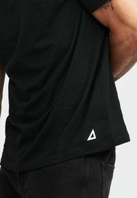 Svart t-shirt i mjuk bomull, med en triangulär vit logotyp på den nedre sidan, något böjd nederkant och avslappnad passform.