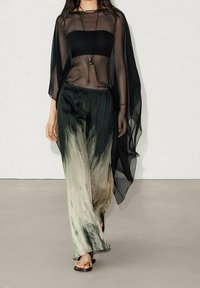 Zwarte doorschijnende top met brede mouwen, gelaagd over een strapless top, gecombineerd met losse, gradient groene en crèmekleurige broeken. Platte sandalen maken de look af.