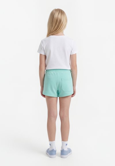 Enfant aux longs cheveux blonds vu de dos, portant un t-shirt blanc, un short vert menthe, des chaussettes blanches et des baskets bleu clair sur fond blanc.