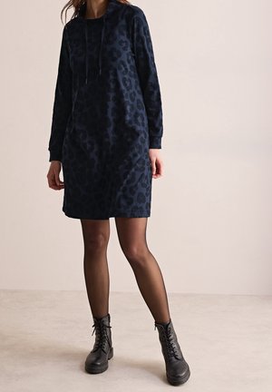Woman wearing a dark blue long-sleeve leopard print dress, sheer black tights, and black lace-up ankle boots, standing on a light floor.