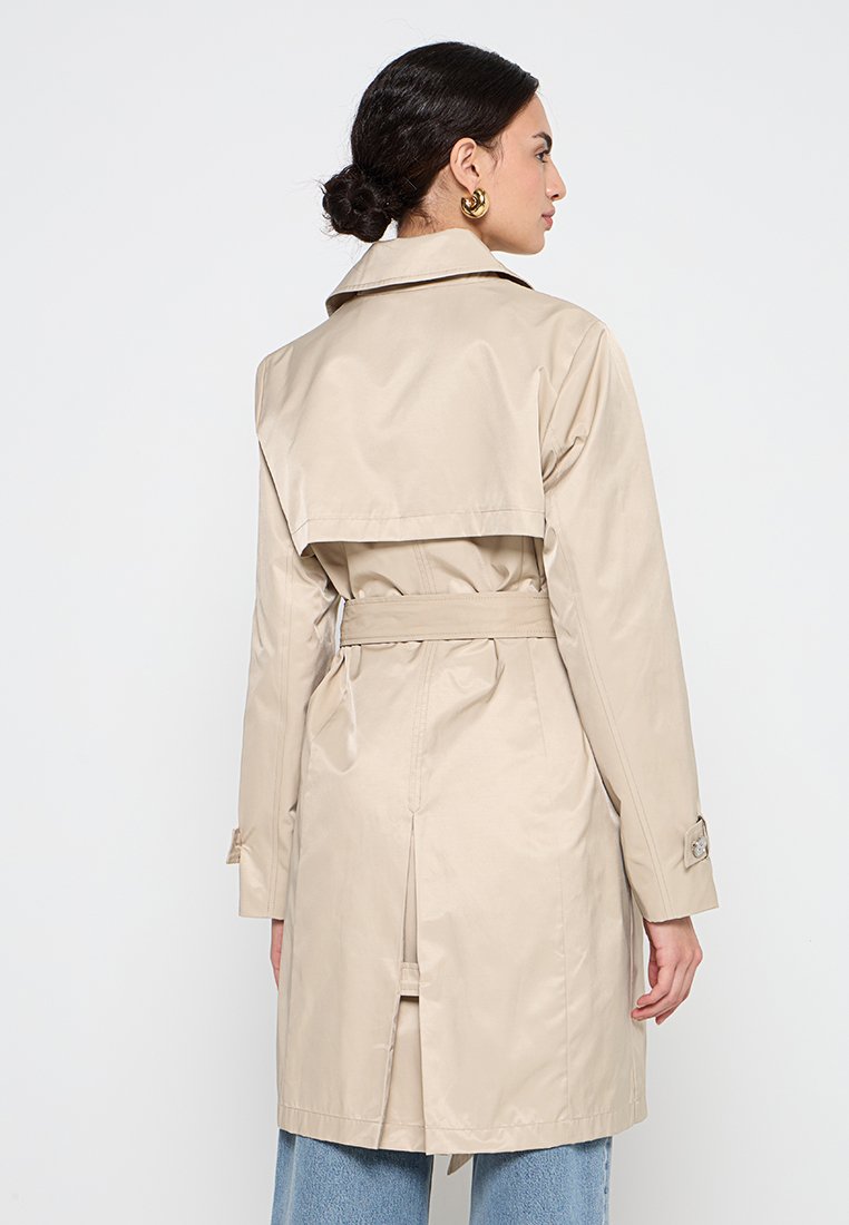 Femme aux cheveux foncés en chignon portant des boucles d'oreilles en or et un trench-coat beige, vue de dos sur fond blanc uni.
