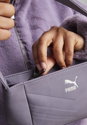 Main aux ongles naturels tenant une tirette de fermeture éclair sur un petit sac Puma violet, porté sur une veste violette en tissu pelucheux avec une fermeture éclair.