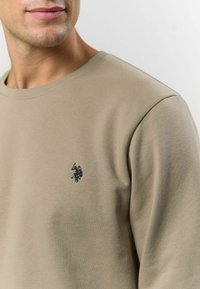 Hanorac bej dintr-un material moale, cu guler rotund și un logo mic brodat în nuanță de bleumarin pe partea stângă a pieptului.