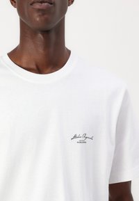 Hvit bomulls T-skjorte med rund hals. Har en svart trykt logo på venstre bryst med teksten "Studio Original Worldwide." Myk tekstur.