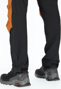 Svarta vandringsskor med texturerat mesh och gummisulor, tillsammans med svarta byxor med orange sidpaneler och dragkedjefickor.