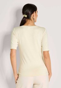 Vrouw in een crèmekleurig shirt met korte mouwen en lichte broek, met haar in een staart gebonden met een gouden haarspeld, met haar rug naar de camera gekeerd.