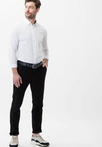 Camicetta bianca con bottoni, pantaloni neri, cintura nera e sneakers con dettagli chiari. Vestibilità moderna, maniche arrotolate e sfondo neutro.