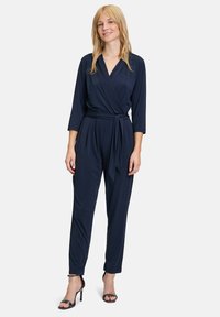 Tuta avvolgente blu navy con maniche lunghe, vita con cintura e gambe affusolate. Realizzata in tessuto liscio, presenta un colletto a V e dettagli minimi.